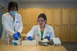 Two students wearing goggles sort metabolic indicator datasets. The students wear lab coats and protective equipment.