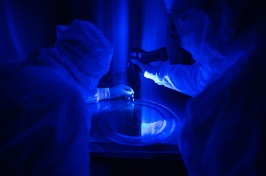 Two people in white Tyvek suits examine a thin flat disk under a blue ultraviolet light that detects dust.
