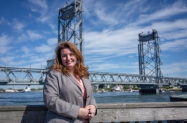 Woman with brown hair wearing gray suit stands in front of Memorial Bridge in Portsmouth.