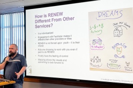 a young white man speaks into a microphone, next to him is a screen with a slide projected on it reading "How is RENEW different from other services?"