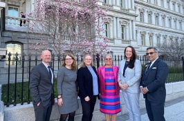 Six people in business attire standing for a photo outside the Eisenhower Executive Office Building in Washington, DC. Behind them is a blooming cherry tree.