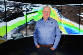 Man in blue shirt stands in front of a screen with mapping graphics on it