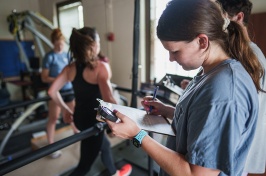 Woman student writes on a clipboard as a runner runs on a treadmill in the background