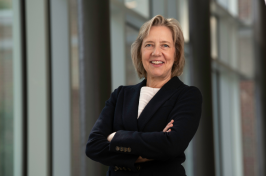 Vanessa Druskat, associate professor of organizational behavior and management at UNH, standing with arms crossed and smiling in a well-lit hallway with large windows.