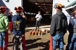 An instructor stands before a group of students, with chainsaws resting on the ground.