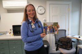 Ruth Swenson stands in a medical office holding a family portrait, wearing a stethoscope and Huggins Hospital ID badge.