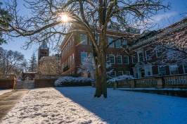 Sunshine glints through tree branches above Thompson Hall, with snowy grass in the foreground