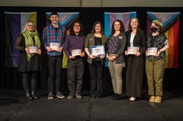 Group of seven award winners pose together on stage holding their awards