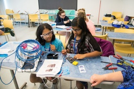 Two young women wearing laboratory glasses sit at a table with cables and equipment to assemble.