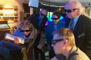 Small group of peope wearing 3D glasses crowd around a computer