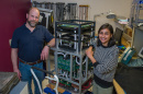 An older man with a beard and a younger woman with dark chin-length hair stand beside a tall cube machine they built. 