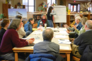 A trained facilitator writes on a large piece of paper while another facilitator leads a discussion at a table of nine participants.