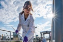 Woman in white lab coat samples liquid outdoors with bright sun over her shoulder