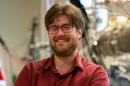 UNH PhD candidate David Ruth smiles at the camera in front of physics equipment