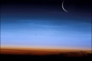 Crescent moon hovers over blue and red sky above the Earth.
