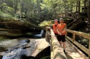 Two men wearing blaze orange vests stand on a log bridge above a stream