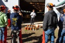 An instructor stands before a group of students, with chainsaws resting on the ground.
