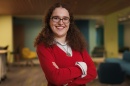 Woman student in red sweater smiles and stands with arms crossed in front of her